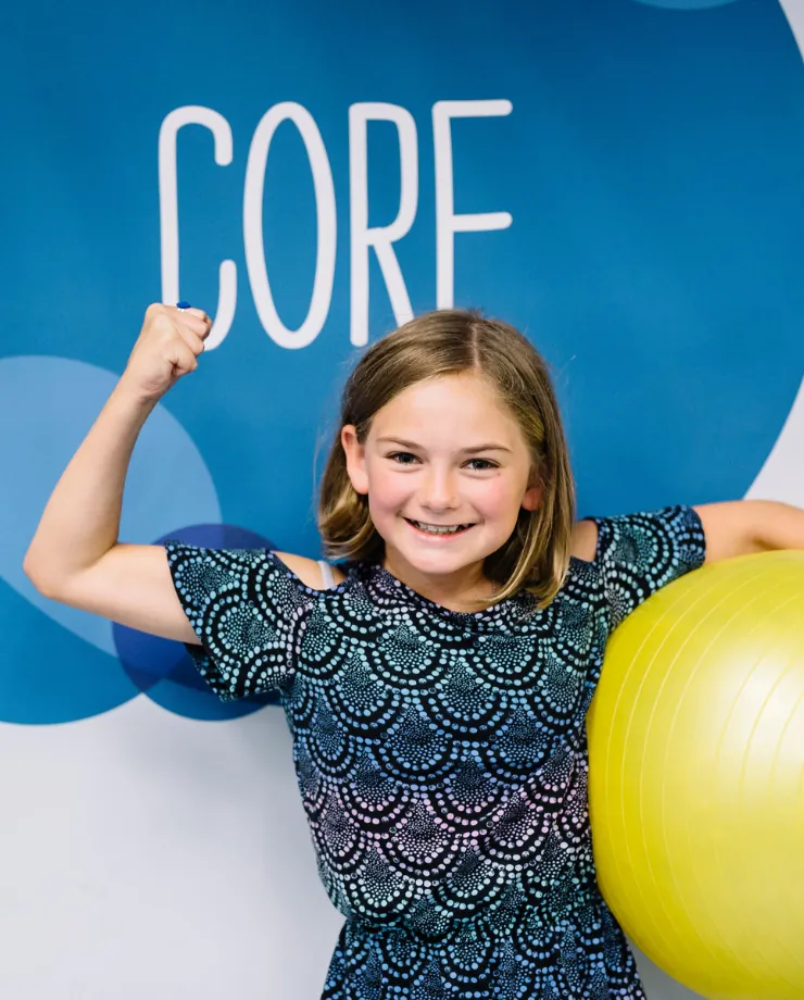 Smiling girl flexing her arm beside an exercise ball in front of a blue wall that reads CORE