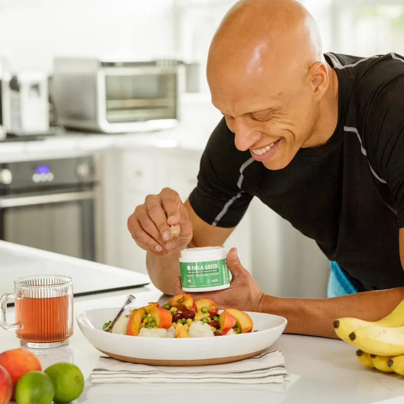 Smiling man sprinkling Amla Green powder over a bowl of fresh fruit in a bright kitchen