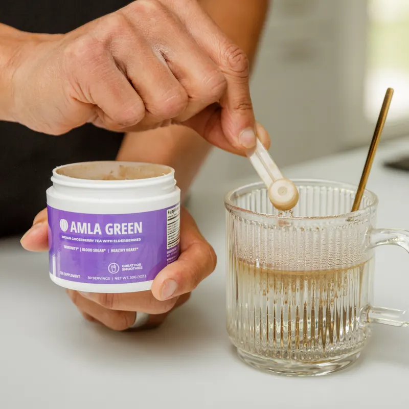 Hand scooping powder from a purple Amla Green jar into a glass mug