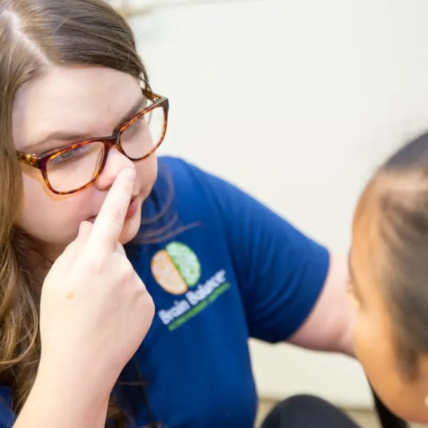 Coach in a Brain Balance shirt touching her nose while demonstrating an exercise for a student